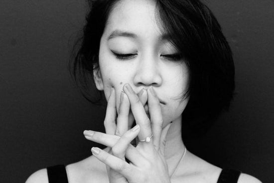 Image for article on how to tell someone you have cancer. Black and white portrait of an East Asian woman. She is looking downwards deep in thought, her hands covering her mouth.