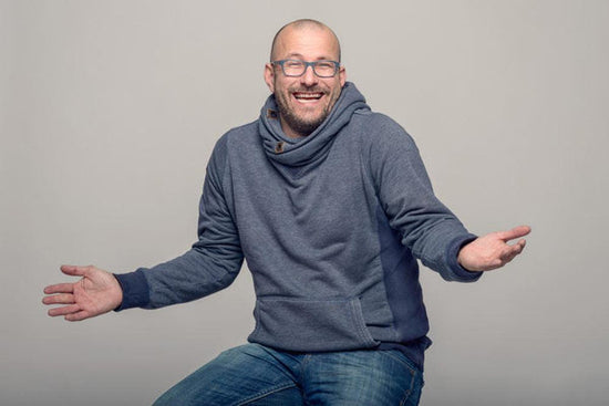 Laughter and cancer: a charismatic balding middle-aged person is wearing bright blue glasses laughing and shrugging as if to say 
