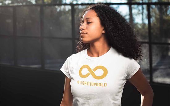 This is a photo of a Black person with an afro wearing a white Light It Up Gold t-shirt. The tee has the word #LightItUpGold printed in large upper case letters beneath a solid line infinity symbol. The symbol and text are colored gold.