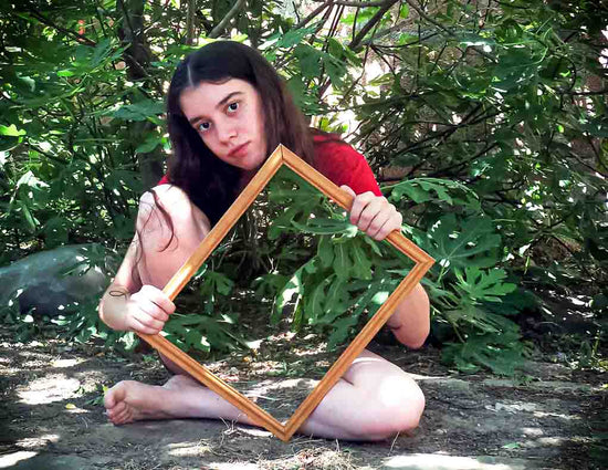 A young woman, living with an invisible disability is sitting cross-legged in a garden. She is holding a mirror in front of her, which makes her torso invisible as we can only see the reflection of the garden.