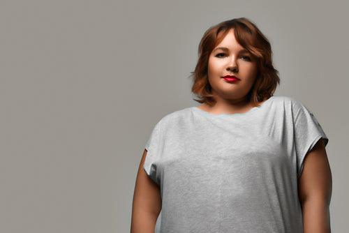 A plus-size / fiercely real/curvy woman with red hair cut in short bob looks intently at the camera. She seems slightly fed-up behind her subtly cheeky closed smile. She wears a grey t-shirt and is standing in front of a grey background.