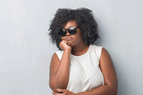 Photo for essay: summer depression is real. A Black woman has her arms crossed, wearing sunglasses, standing against a grey wall. 