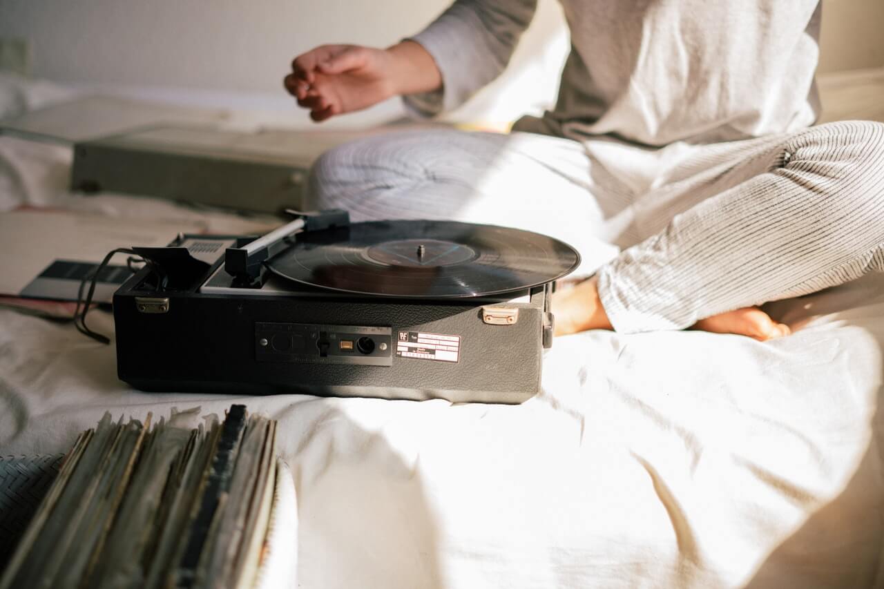 person seated on bed with white sheets listening to an album on a record player