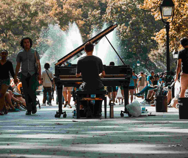 Musician playing piano on a busy street with fountains and trees in the background.
