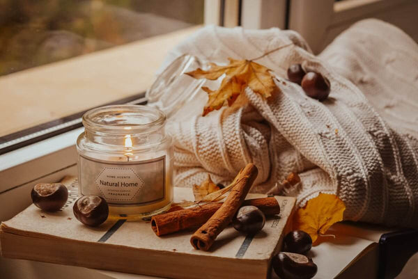 A scented candle next to a white blanket, cinnamon sticks, and chestnuts