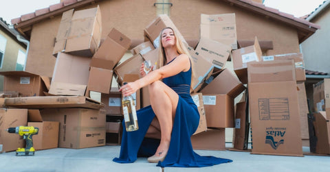 Woman wearing a blue dress with a bottle of champagne in her hand squatting in front of a house and a pile of moving boxes