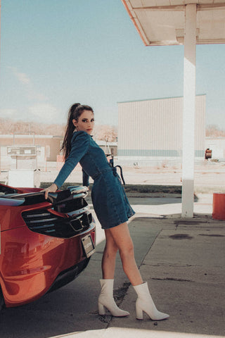 Woman next to a sports car wearing denim dress and white ankle boots