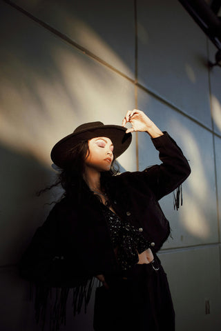 Woman posing on a wall wearing a fringed jacket and a wide-brimmed hat