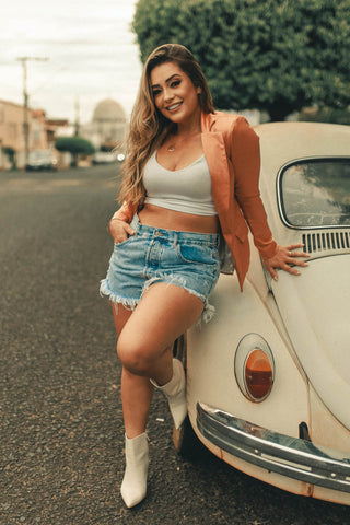 Woman posing for a photo next to a retro car wearing denim shorts and a crop top with blazer