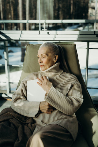 Elderly woman sitting on a sofa and holding a book wearing a bulky turtleneck