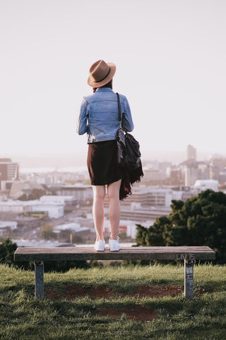 Photo of the back of a woman standing on a bench and wearing a jeans jacket and brown skirt with sneakers