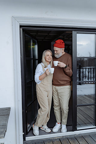 Elderly couple posing at the door and holding coffee cups wearing casual outfits