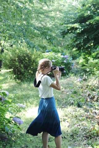 Woman in a forest taking a photo wearing a blue skirt and a shite t-shirt
