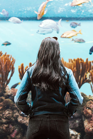 Frau steht vor einem Aquarium und trägt blaue Jeansjacke und graue Jeans