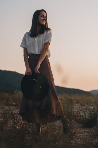 Femme posant dans des champs ouverts avec une longue jupe marron, un bouton blanc et tenant un chapeau à larges bords