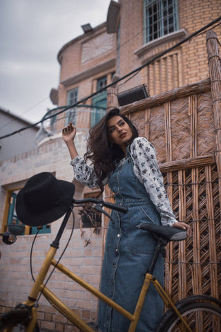 Young woman posing next to a bicycle and wearing a denim skirt romper and a floral shirt