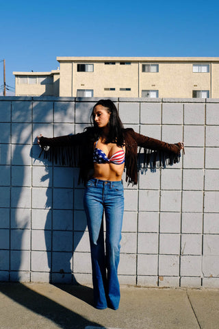 Woman laying on a wall posing for a photo wearing a USA bralette top, flared jeans and a fringed jacket