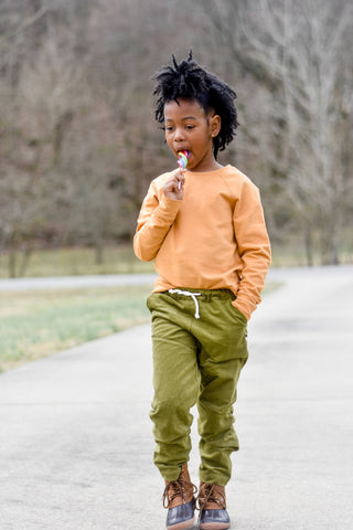 Young child in orange sweatshirt and green pants, walking while licking a lollipop