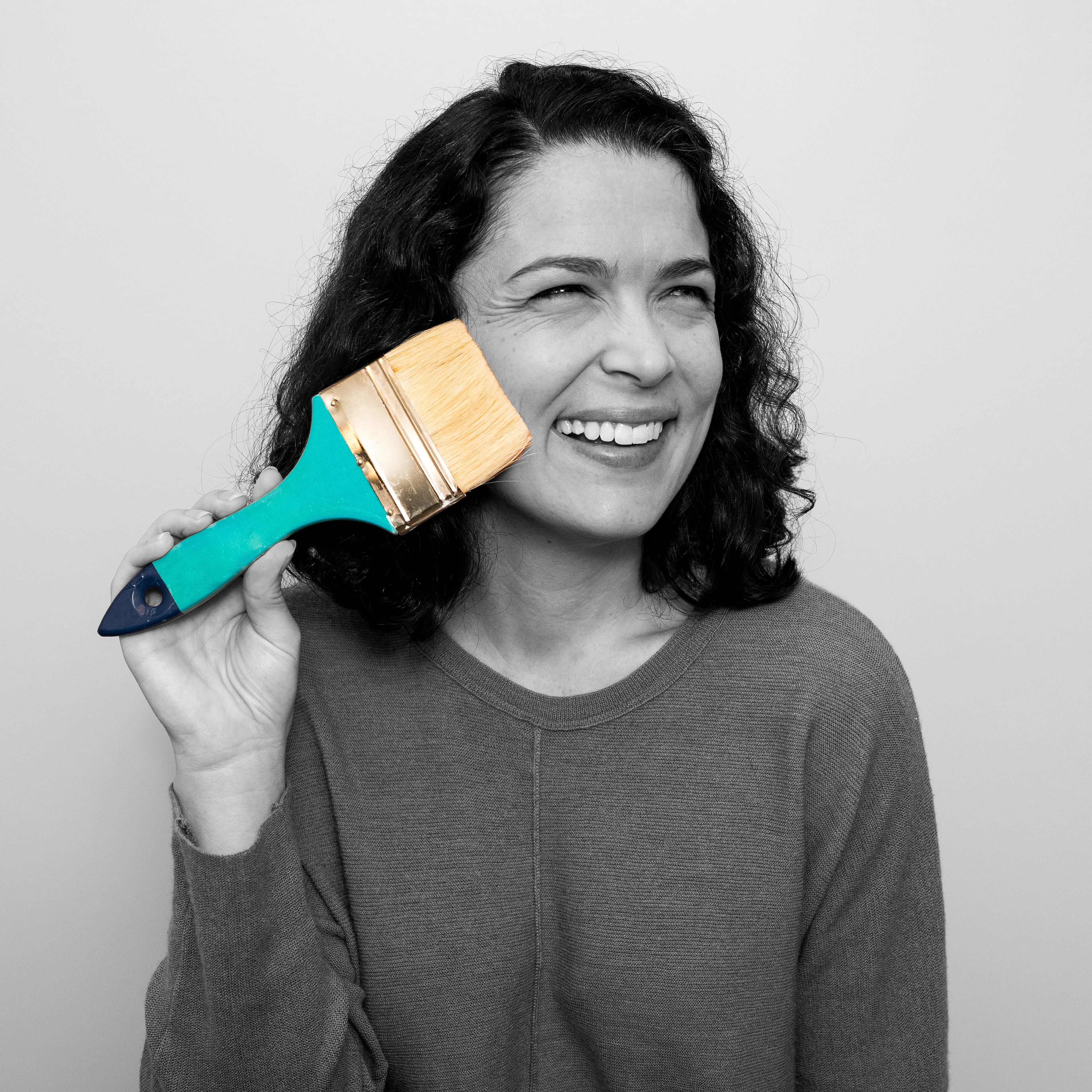 Black and white portrait of a woman with curly shoulder length hair smiling, and holding a teal paintbrush to her face