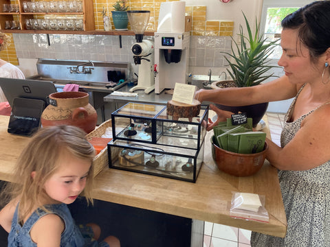 picture of a woman placing jewelry into a glass case on a cafe counter while a small child sits at the counter