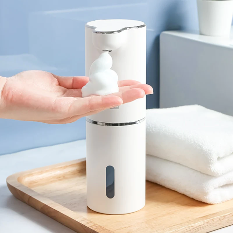 Una mano, saca jabón de espuma de un dispensador blanco, que esta sobre una bandeja de madera. Al lado del dispensador hay dos toallas blancas.