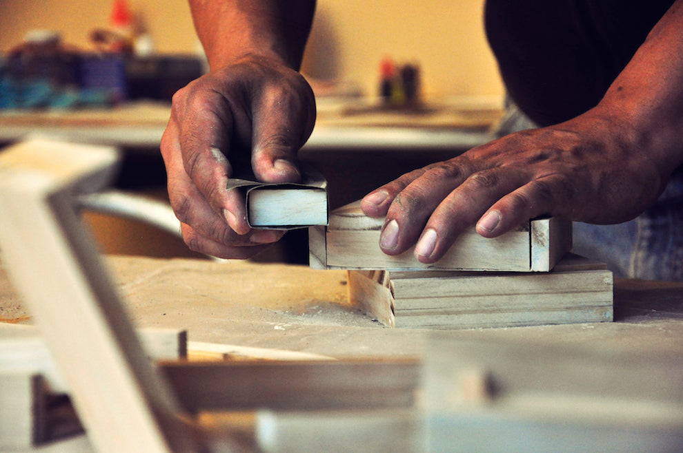 Close-up photograph of someone handcrafting a wooden box on a table. Their hands are dusty and worn, and they are using sandpaper to smoothen the edges of the box. There is sawdust on the table, and in the background are tools.