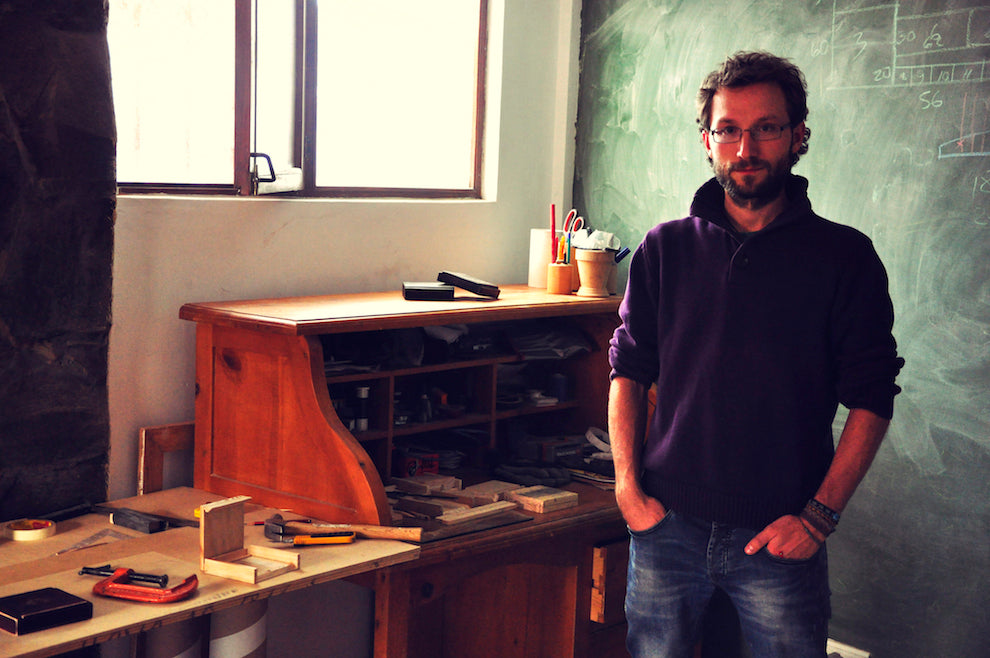 Photograph of To’ak’s co-founder, Carl Schweizer. He is standing in the middle of a studio, with tools and crafted wood boxes on the table beside him. In the background is a blackboard with notes written in chalk.