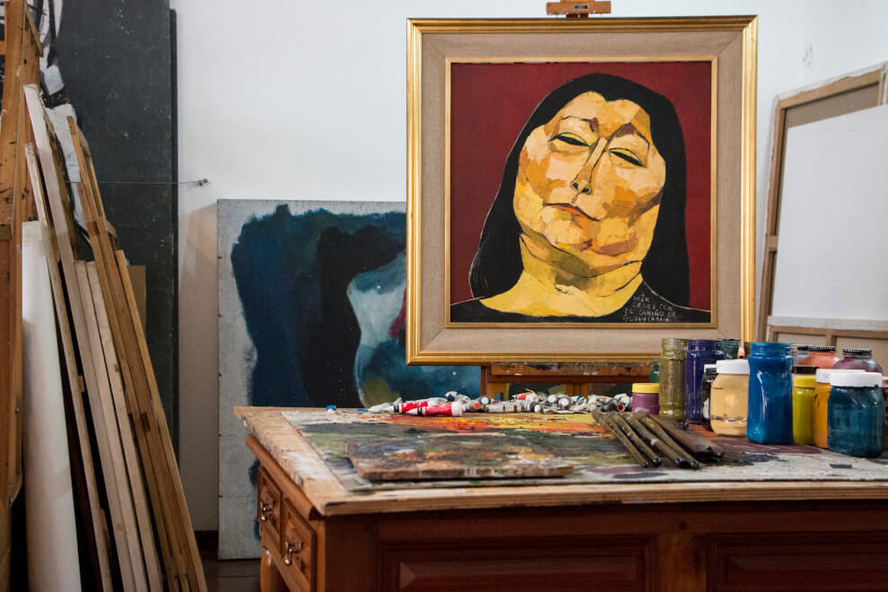 A painting by artist Oswaldo Guayasamín depicting a dark haired woman sits on a desk with paints