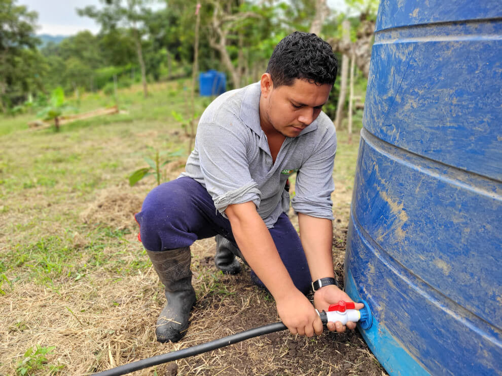 Mario kneels as he connects a rubber hose to the bottom of a blue vat (irrigation system)