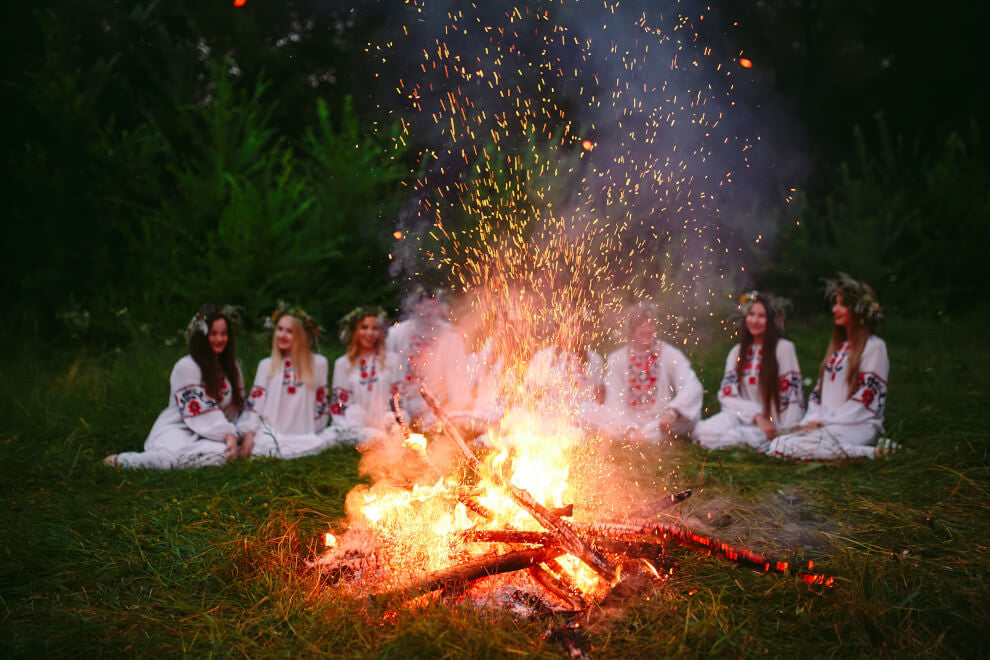 eight women sit in white dresses with flower crowns in front of a bonfire
