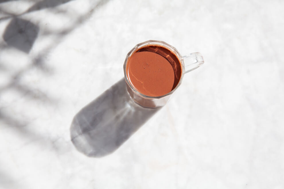 Pictured from above: a cup of hot chocolate sits on a white marbled surface. In the corner is the shadow of a tree's leaves and this branches