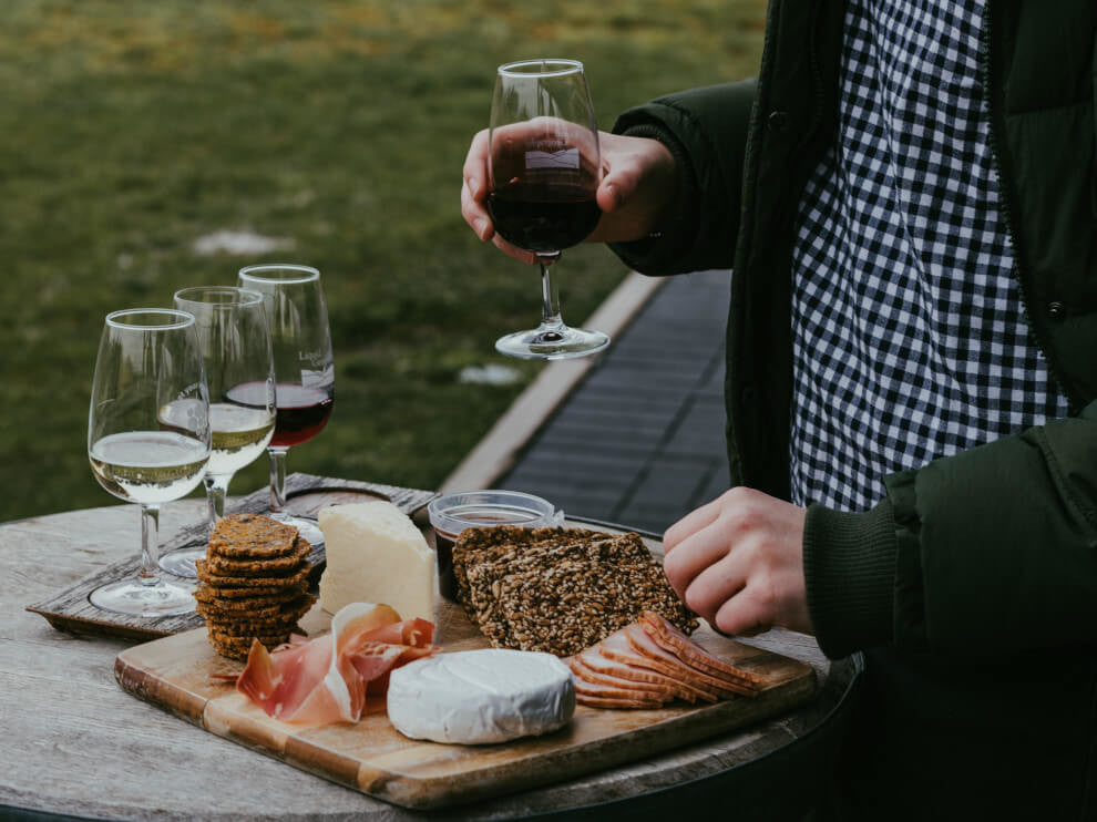 a man stands, wine glass in hand, in front of a cheese and meat board 