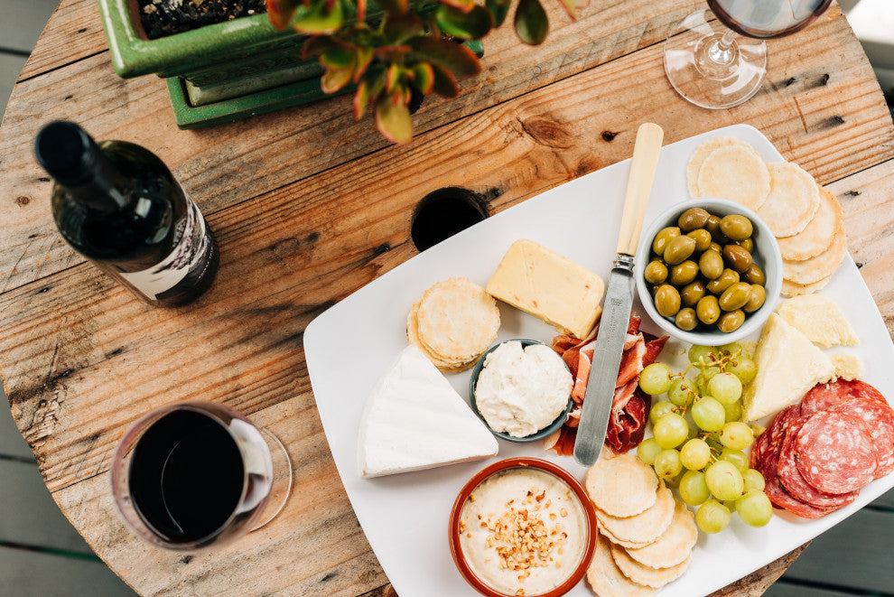 A platter of cheeses, fruits, and sliced meats sit on a wooden table beside a glass of red wine