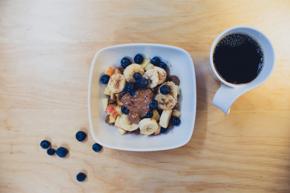 a cup of coffee alongside a plate of fruit with cacao powder and peanut butter on top