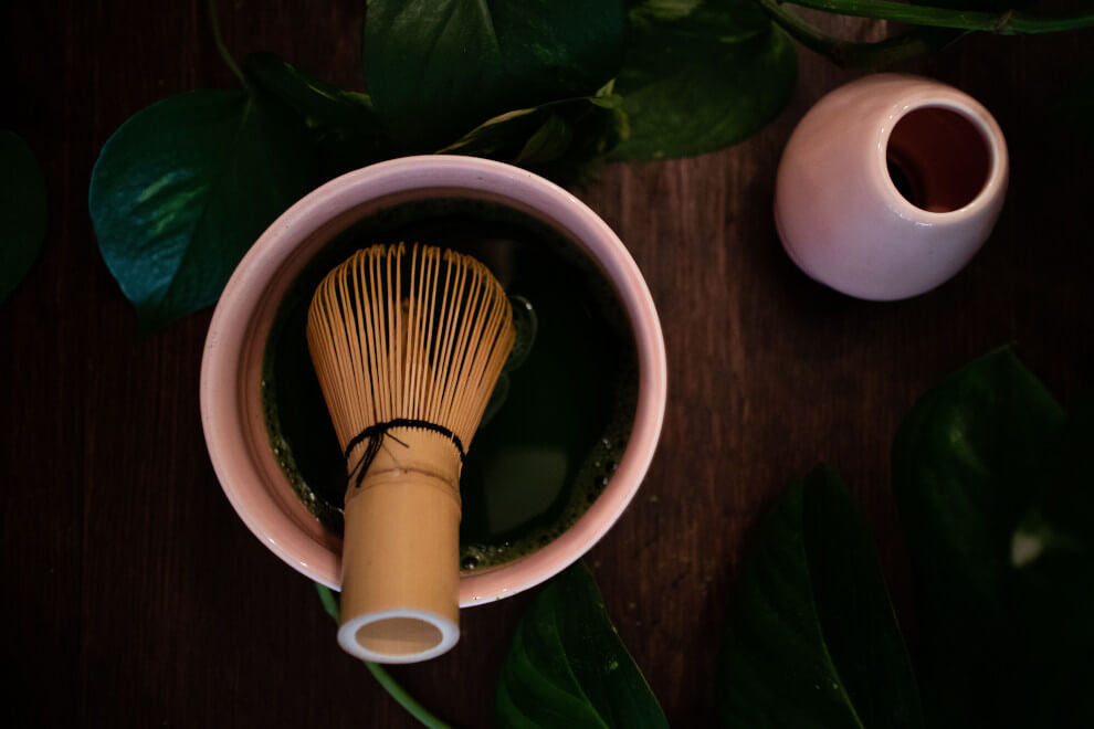 a wooden whisk sits in a pink bowl containing matcha tea