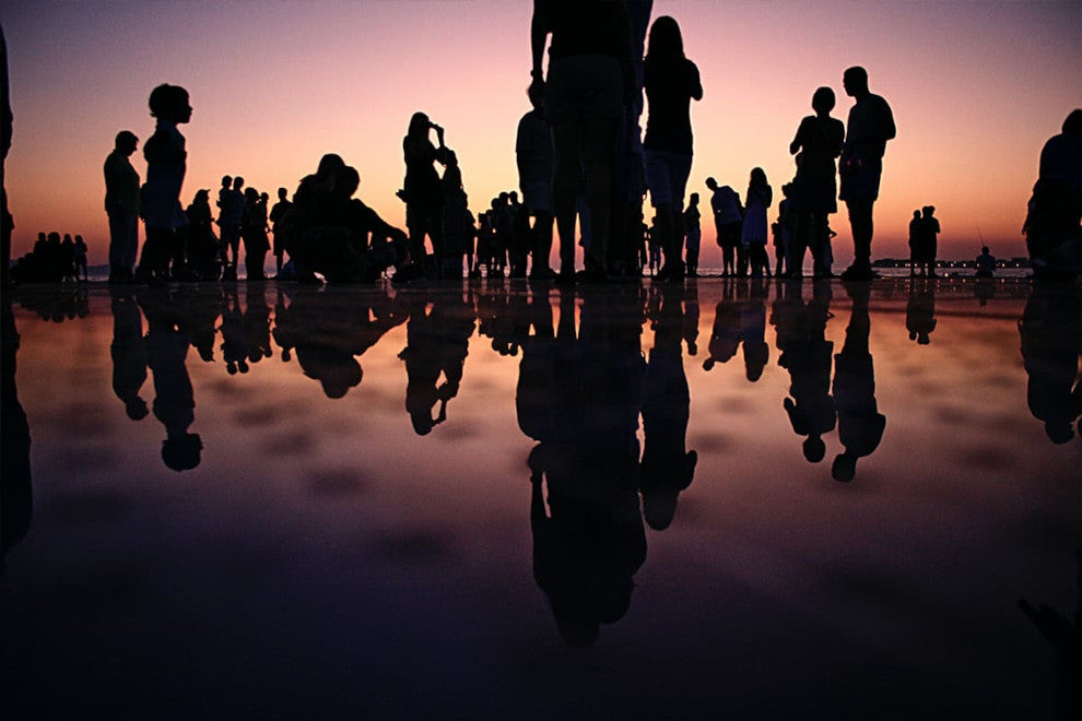 sillhouetes of around 15 people stand in a shallow pool of water. The sun is setting in the backdrop