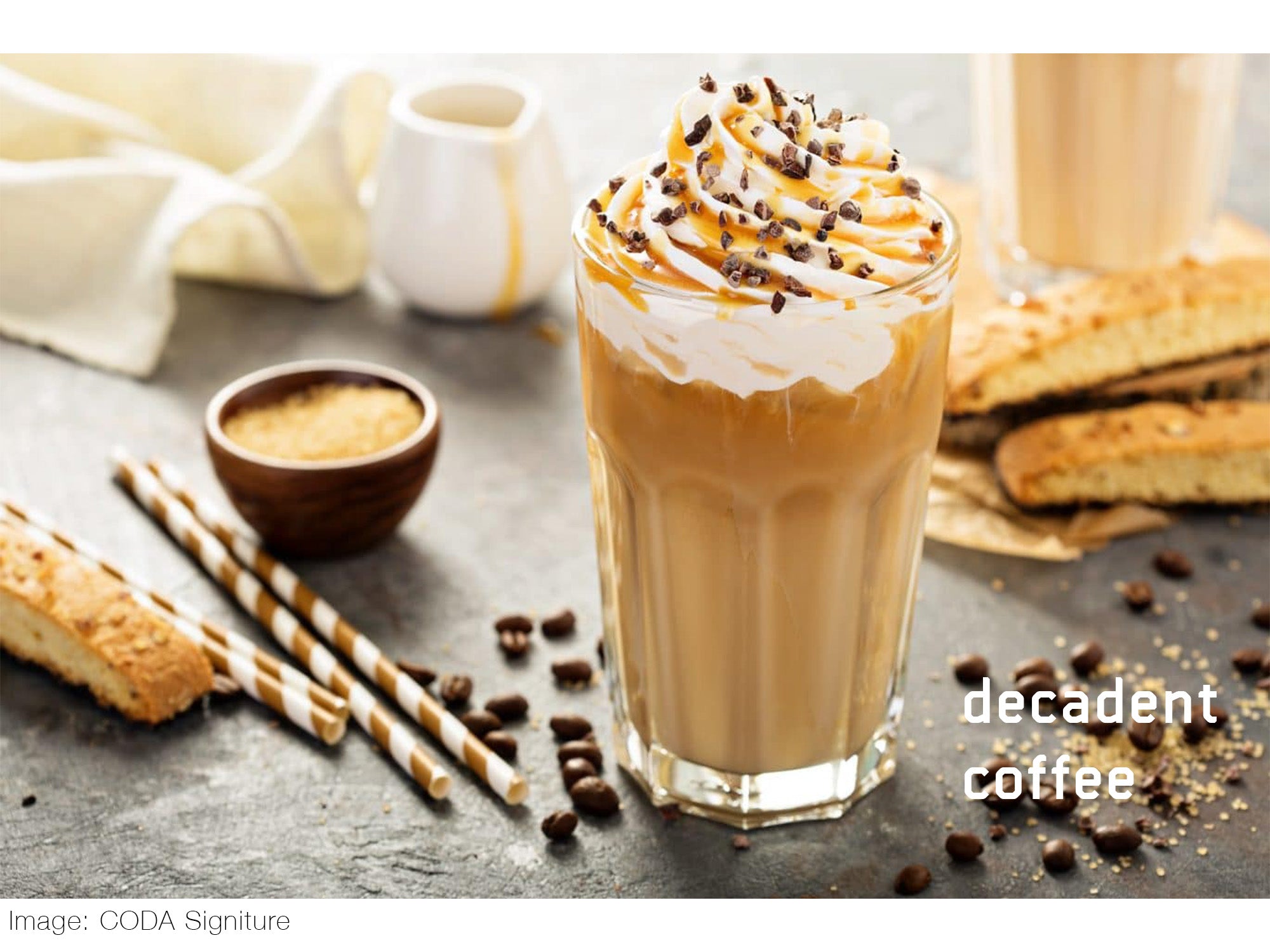 Creamy coffee topped with whipping cream and caramel sauce in a clear cup, on a table with spilled coffee beans, paper straws, and sugar