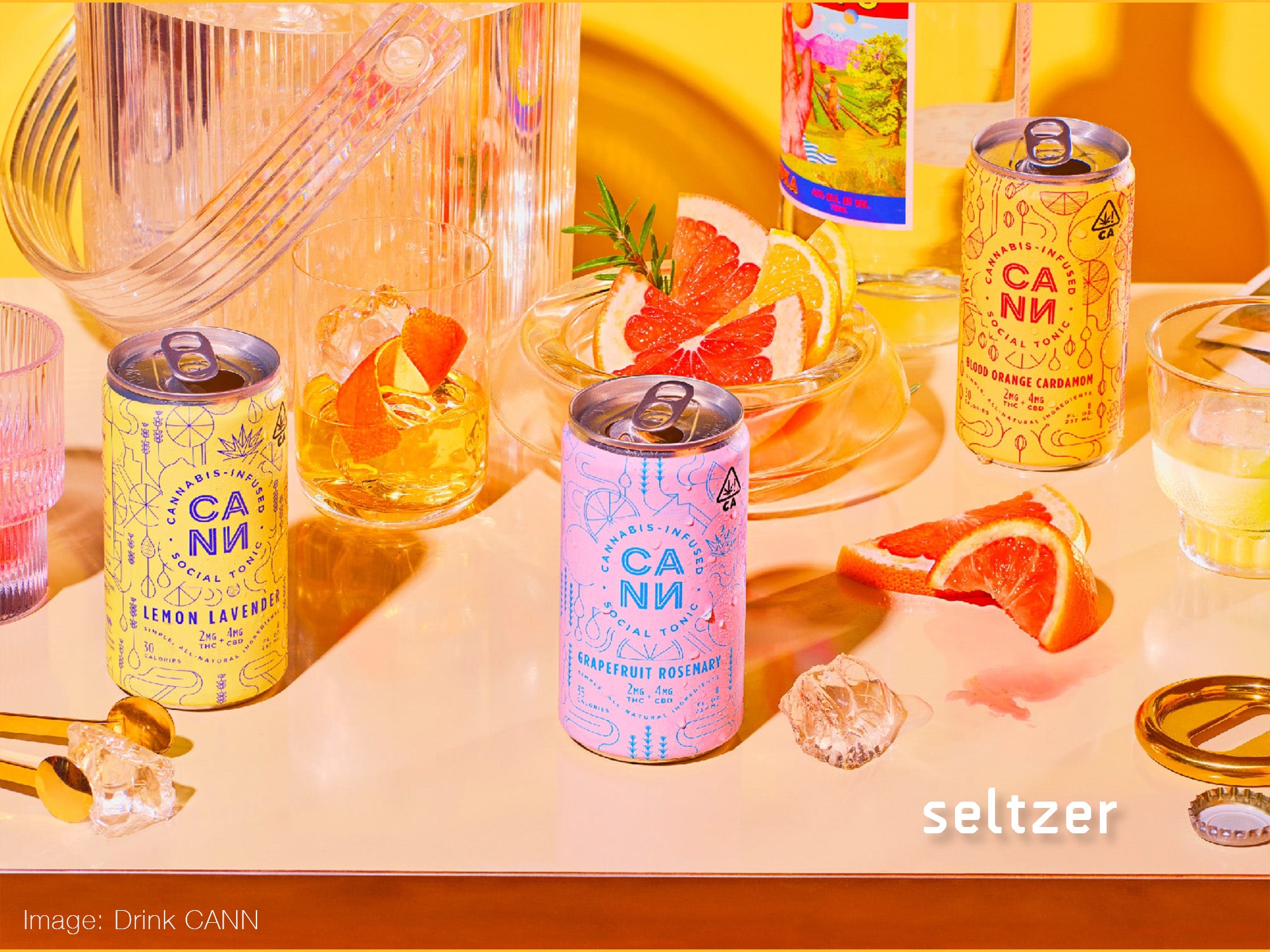 CANN cannbis infused selzters on table scape in three different flavours, surrounded by cocktail glasses, ice, cut fruit, and a festive atmosphere