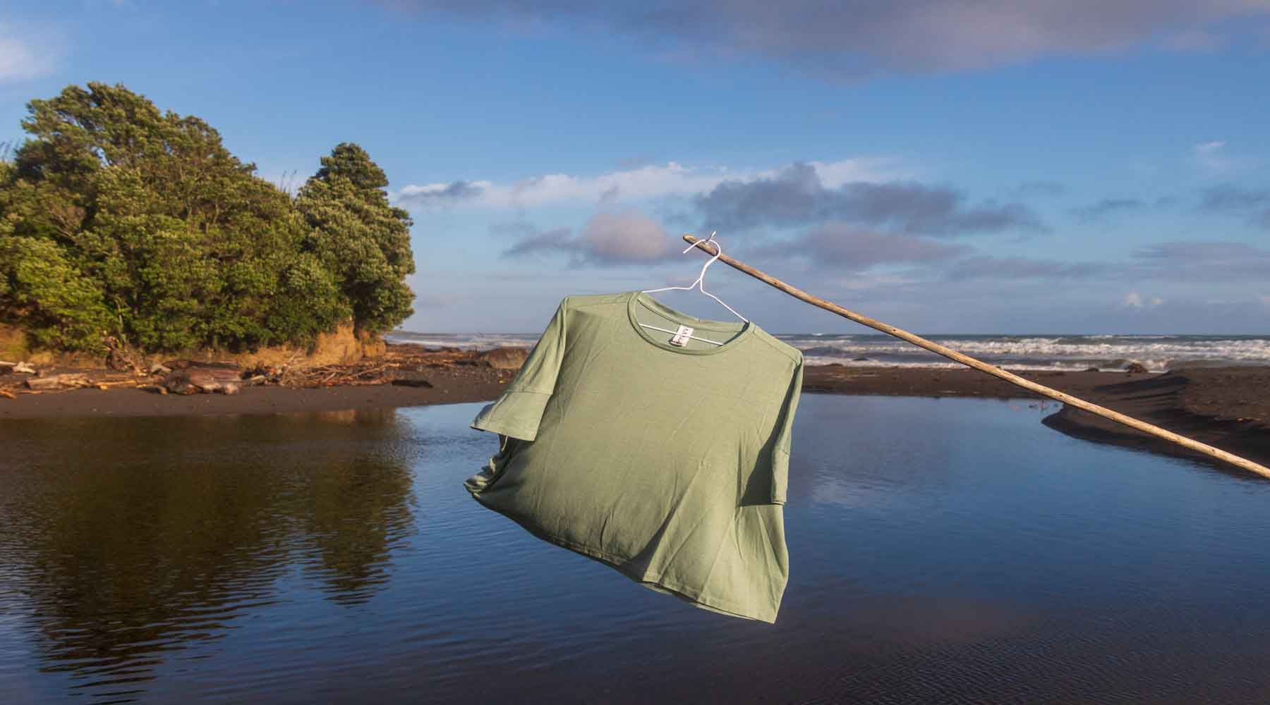 Asmuss Sea Green Aline Tshirt on a coat hanger hanging off a stick above an estuary at the black sand beach of Oakura New Zealand