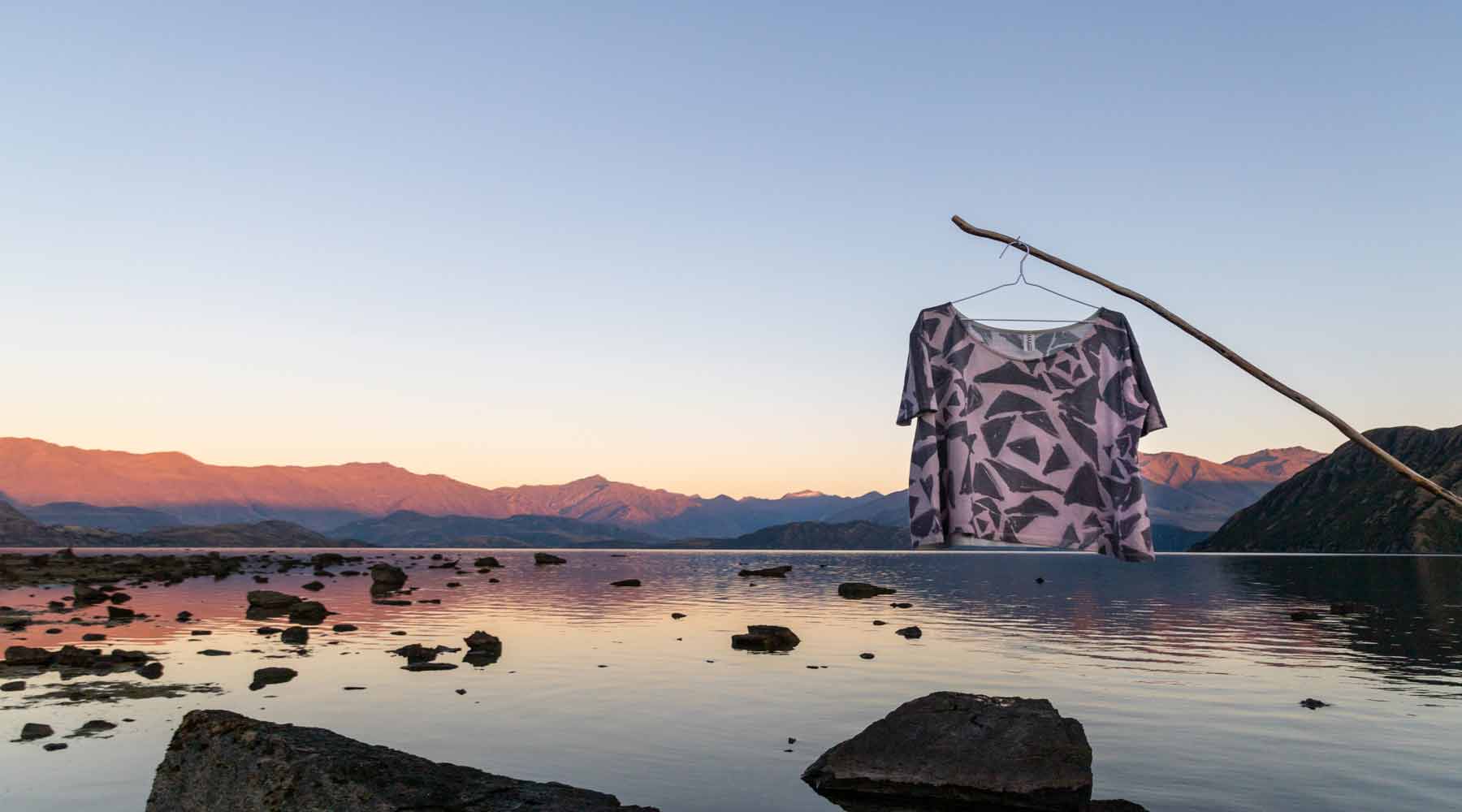 Asmuss Black Rose Tshirt on a coat hanger hung off a stick above the still water and rocks of Lake Wanaka while the sun rises on the mountains in the distance