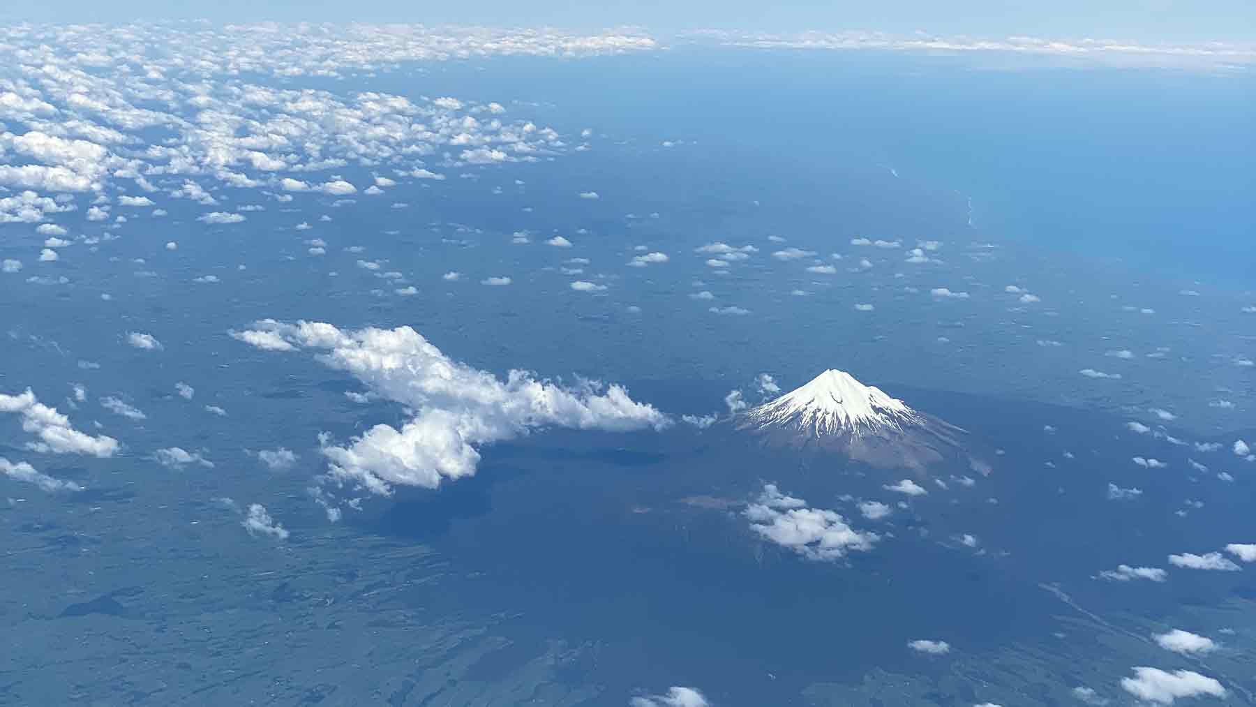 Snow covered Mount Taranaki and the coast of Taranaki from an airplane with little fluffy white clouds