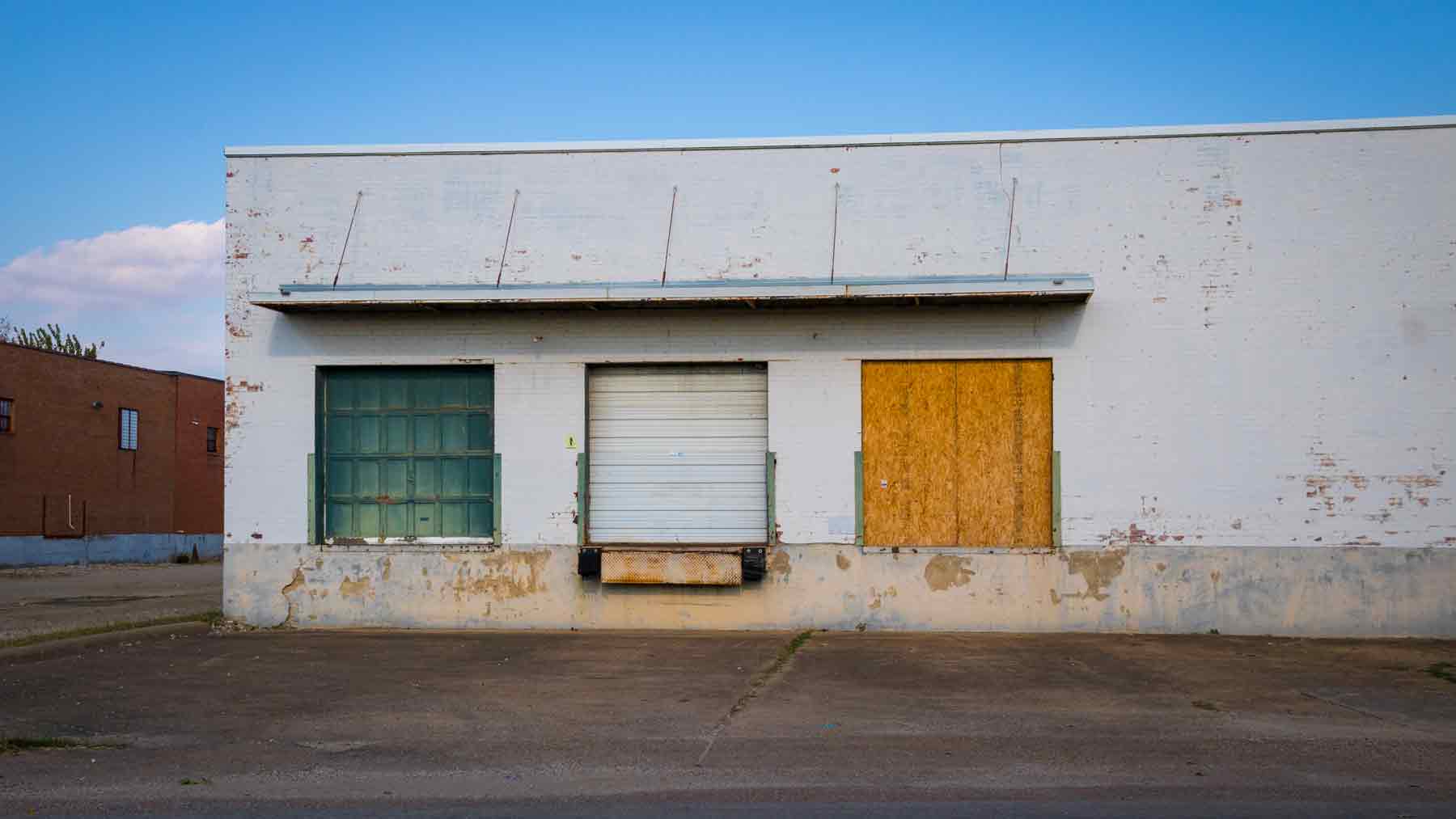 Old white factory loading dock with one green metal door, one white plastic door and one door with chip board