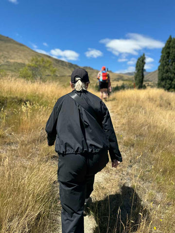 Asmuss cofounder Fiona walking the Fern Burn Track wearing the Asmuss Roam Jacket and Oval Trousers. Her sister Andrea is further ahead and the sky is bright blue with small white clouds