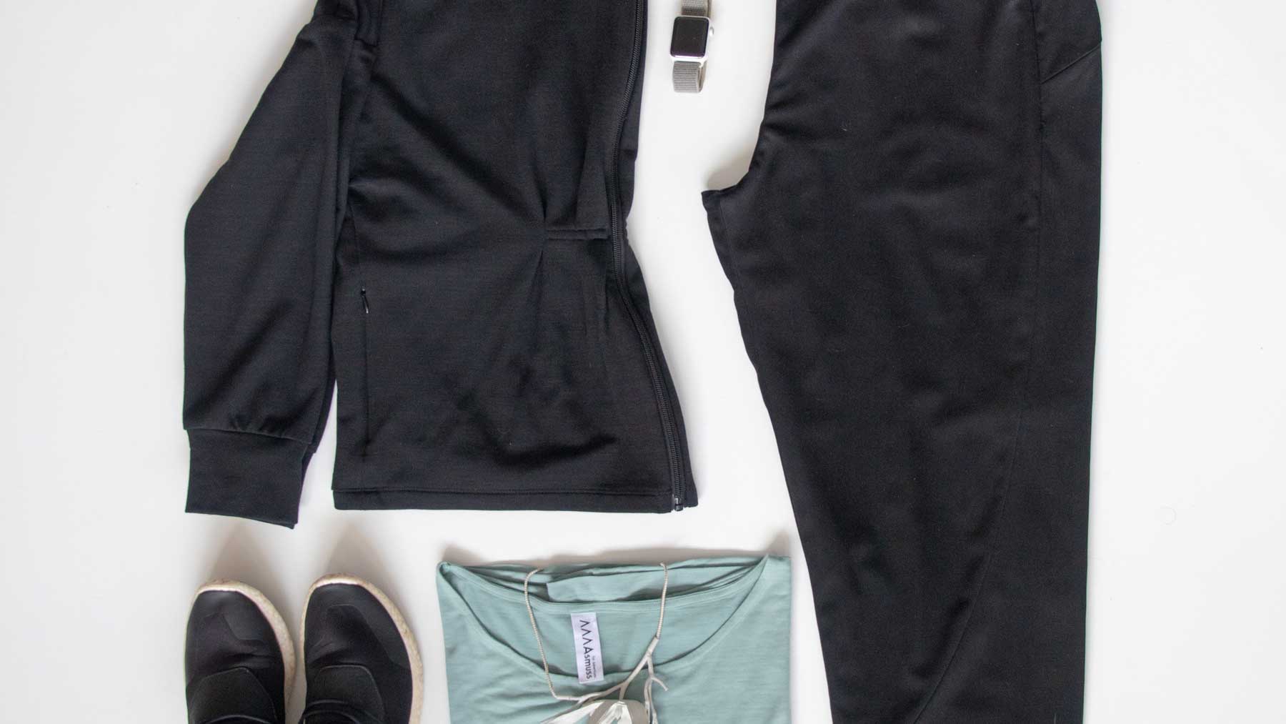 Flat lay image of Asmuss clothing to pack for travel to Fogo Island including the Asmuss Pleated Zip Through Sweat, Asmuss Panelled Trousers and Asmuss A-line T-shirt in sea green along with accessories to finish a great travel outfit off