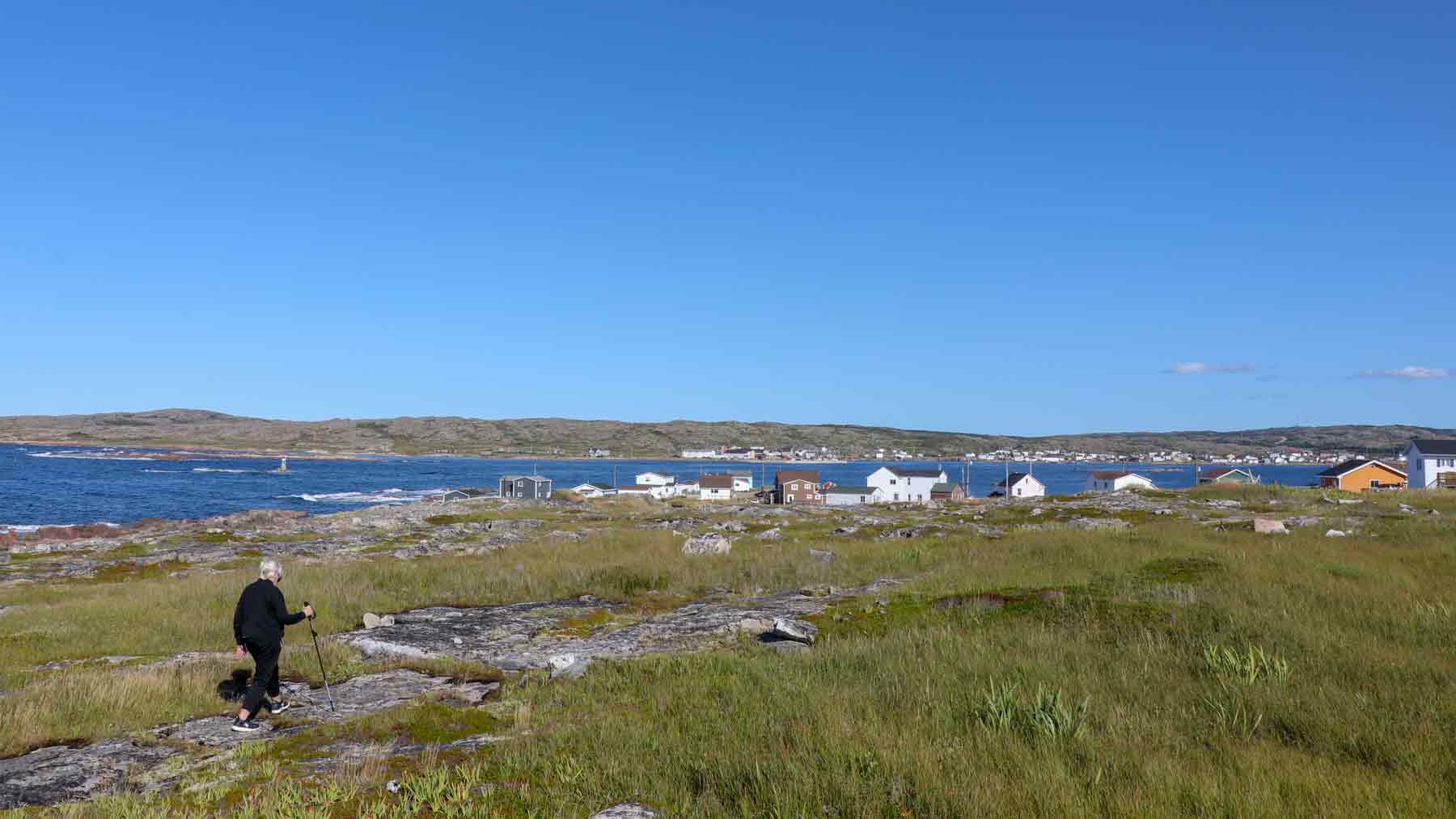 The Asmuss founder's Mum walking on Fogo Island with cottages in the distance on the coast. She is wearing the Asmuss Pleated Zip Through Sweat and Asmuss Panelled Trousers and looks great