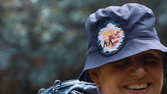 Fiona, an Asmuss co-founder, wearing a sample of the  Asmuss bucket hat with Evelyn rose inspired embroidered badge with a backpack on.  She is smiling rather than grimacing.