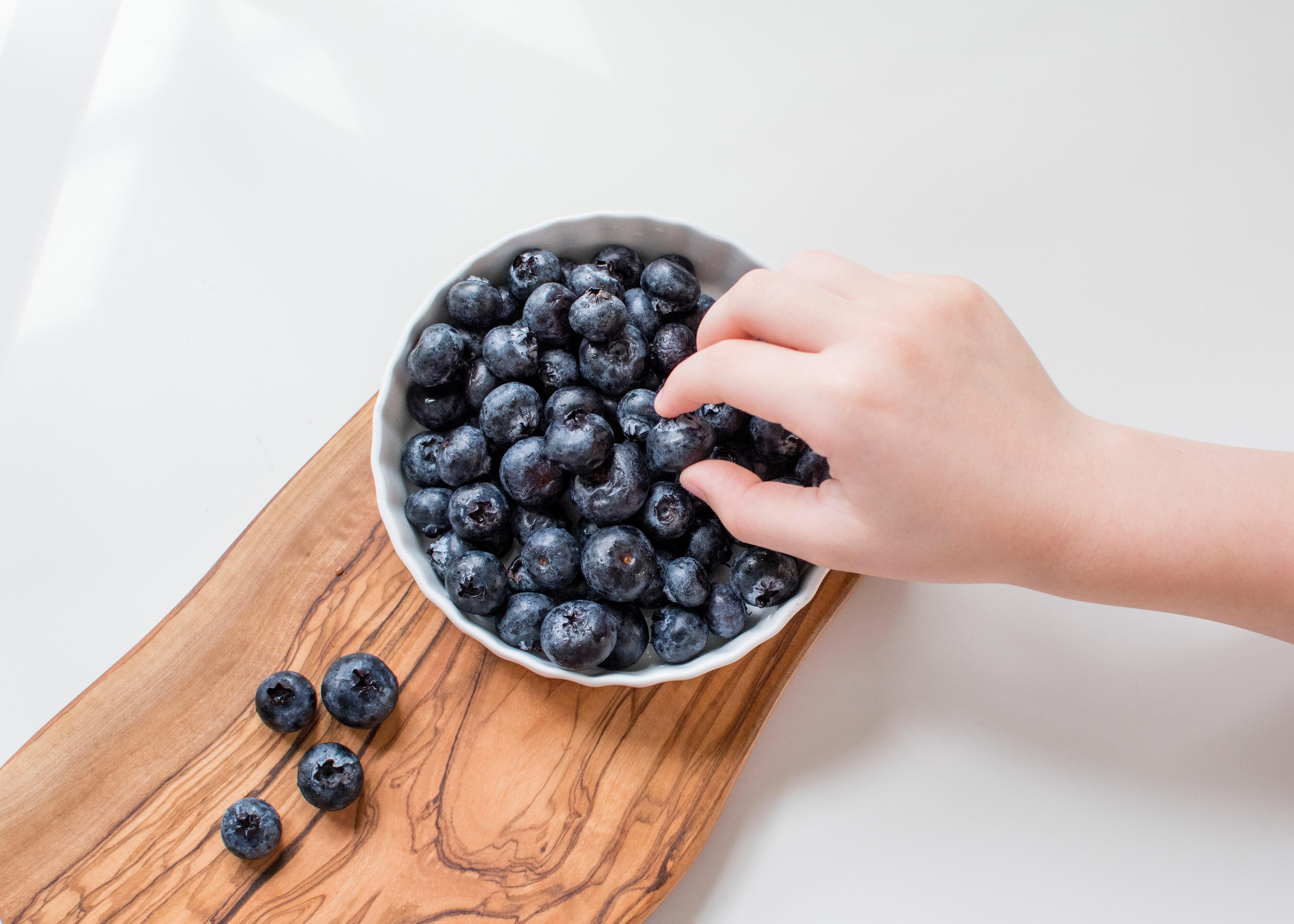 Caucasion hand reaching into white bowl filled with blueberries on a wooden serving board.