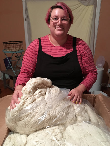 Photo of Mckenzie with a wary smile on her face standing over a massive box containing many skeins of wool.