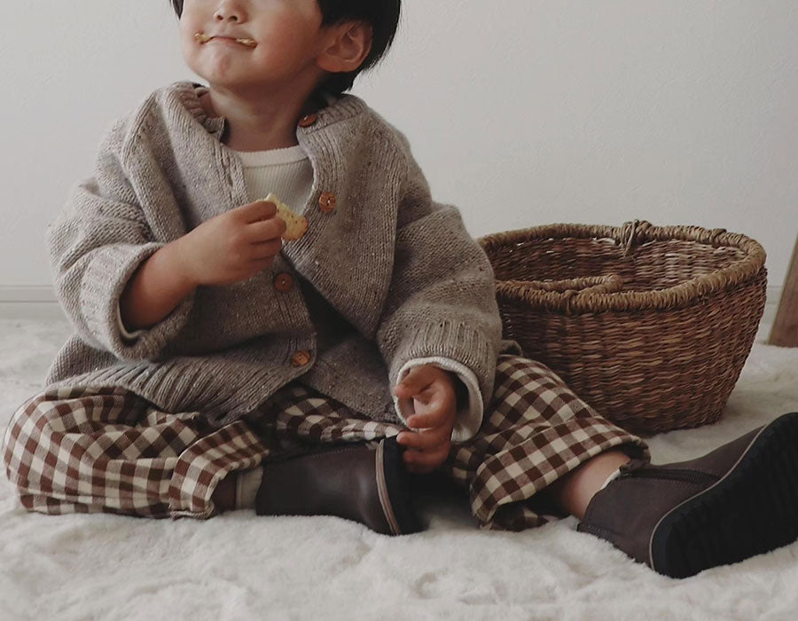 Toddler in oatmeal wool cardigan, brown gingham pants & boots, sitting & eating biscuits