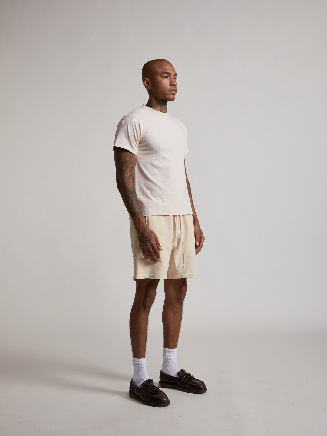 bald man with tattoos wearing bare knuckles fitted crew neck t shirt in smoke white with lounge shorts and loafers with socks.